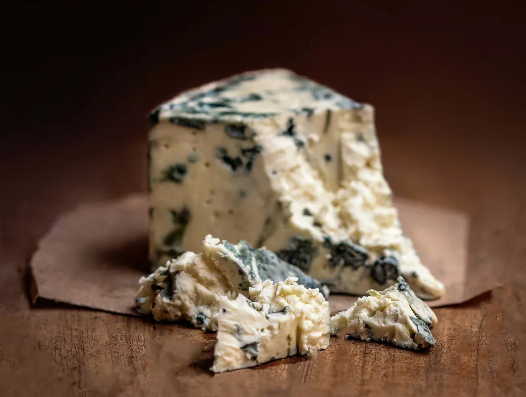 Nahaufnahme von Blauschimmelkäse mit blauer Aderung auf einer Holzunterlage. Ein großes Stück ist von zerbröselten Stücken umgeben.