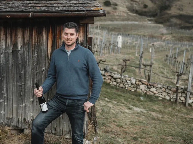 Ein Mann in einem blauen Pullover und Jeans steht mit einer Weinflasche in der Hand neben einem rustikalen Holzschuppen in einem Weinberg.
