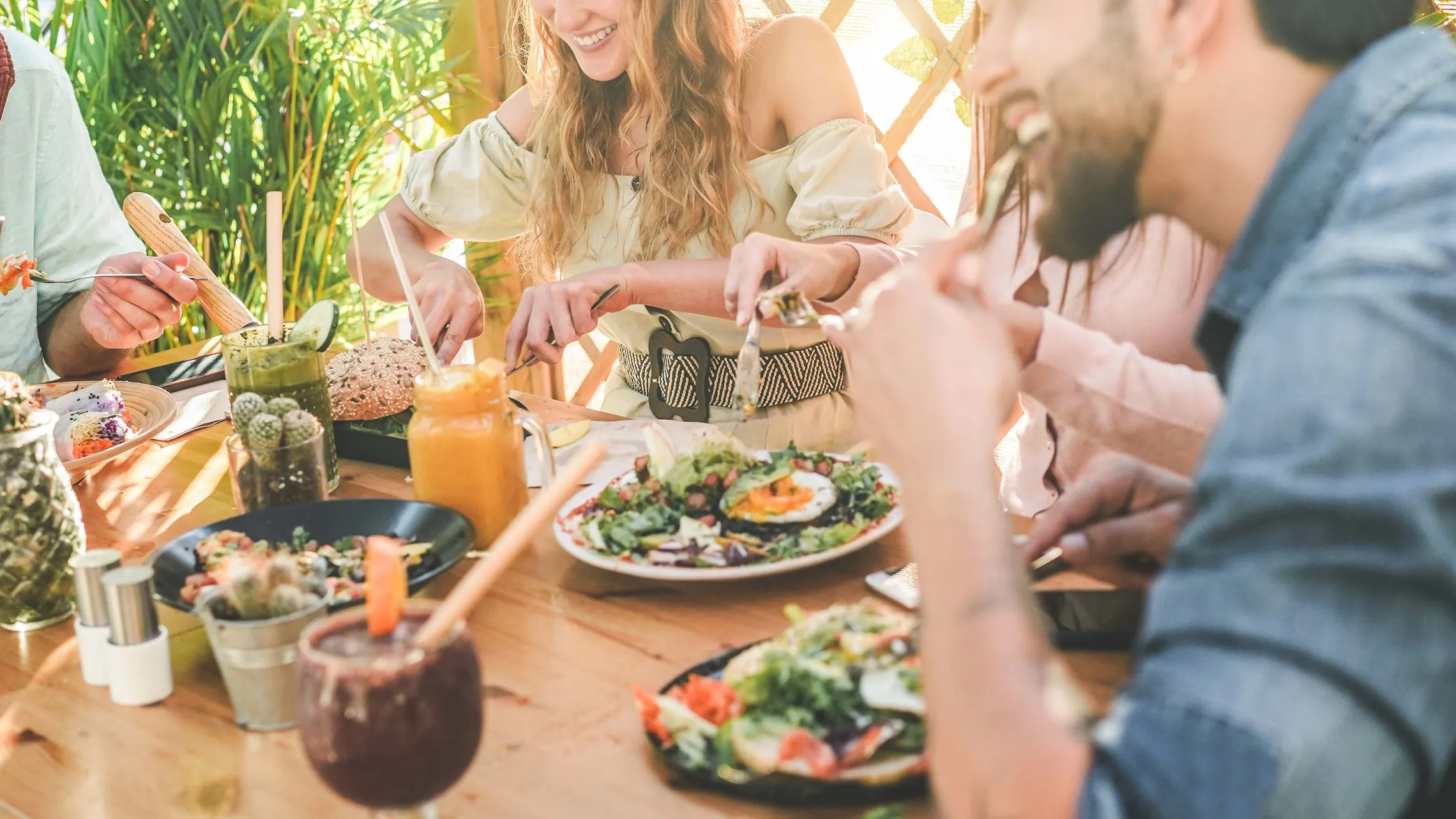 Eine Gruppe genießt an einem Tisch im Freien eine Mahlzeit mit Salaten und Smoothies, umgeben von Grün und Sonnenschein.