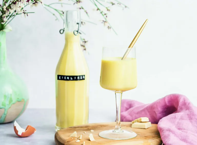 Eine Flasche und ein Glas gelber Eierlikör mit der Aufschrift „Eierlikör“ auf einem Holzbrett mit Eierschalen und Schokoladenstückchen, daneben ein rosa Tuch.