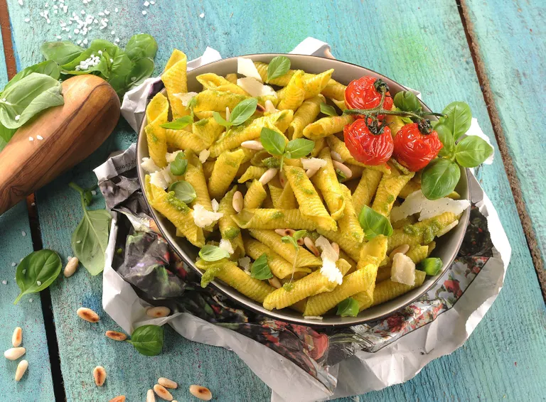 Eine Schüssel Pasta mit Pesto, garniert mit Kirschtomaten, Basilikum, Pinienkernen und Käsespänen, auf einem rustikalen Holztisch.