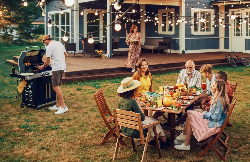 Eine Gruppe von Menschen genießt ein Barbecue im Freien mit Lichterketten; sie sitzen um einen Tisch herum, der mit Essen gedeckt ist, während ein Mann in der Nähe grillt.