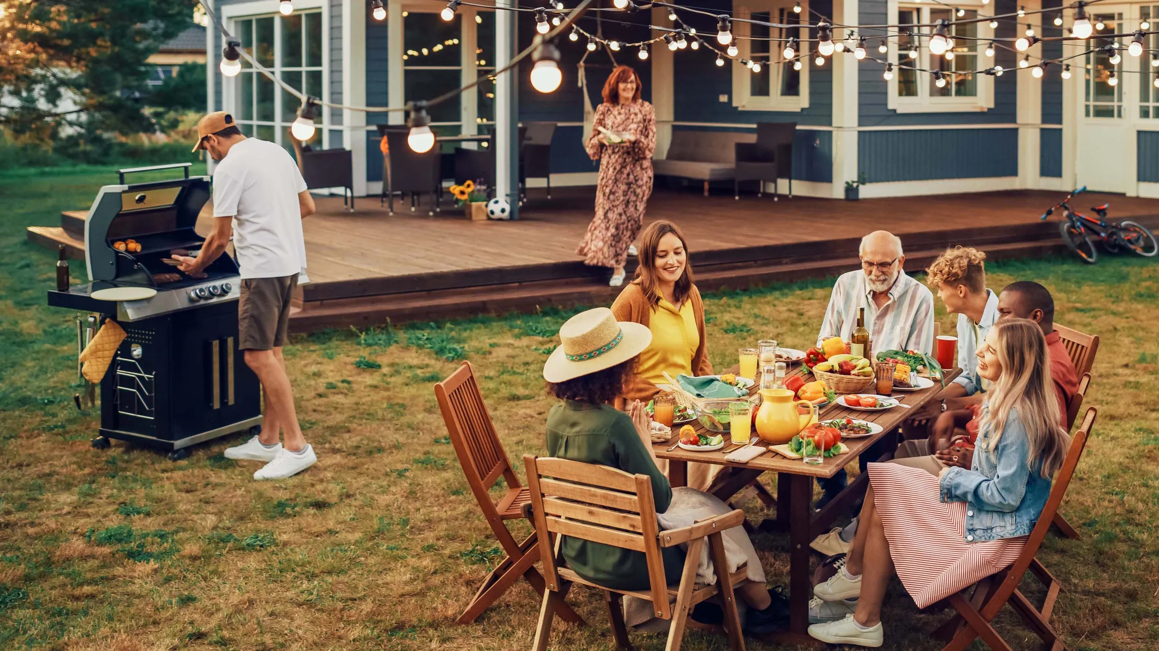 Eine Gruppe von Menschen genießt ein Barbecue im Freien mit Lichterketten; sie sitzen um einen Tisch herum, der mit Essen gedeckt ist, während ein Mann in der Nähe grillt.