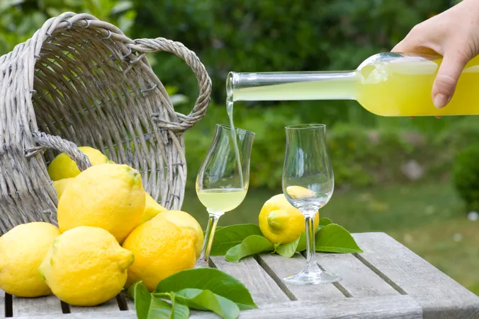Eine Hand gießt Zitronenlikör aus einer Flasche in ein Glas, umgeben von frischen Zitronen und Blättern auf einem Holztisch.