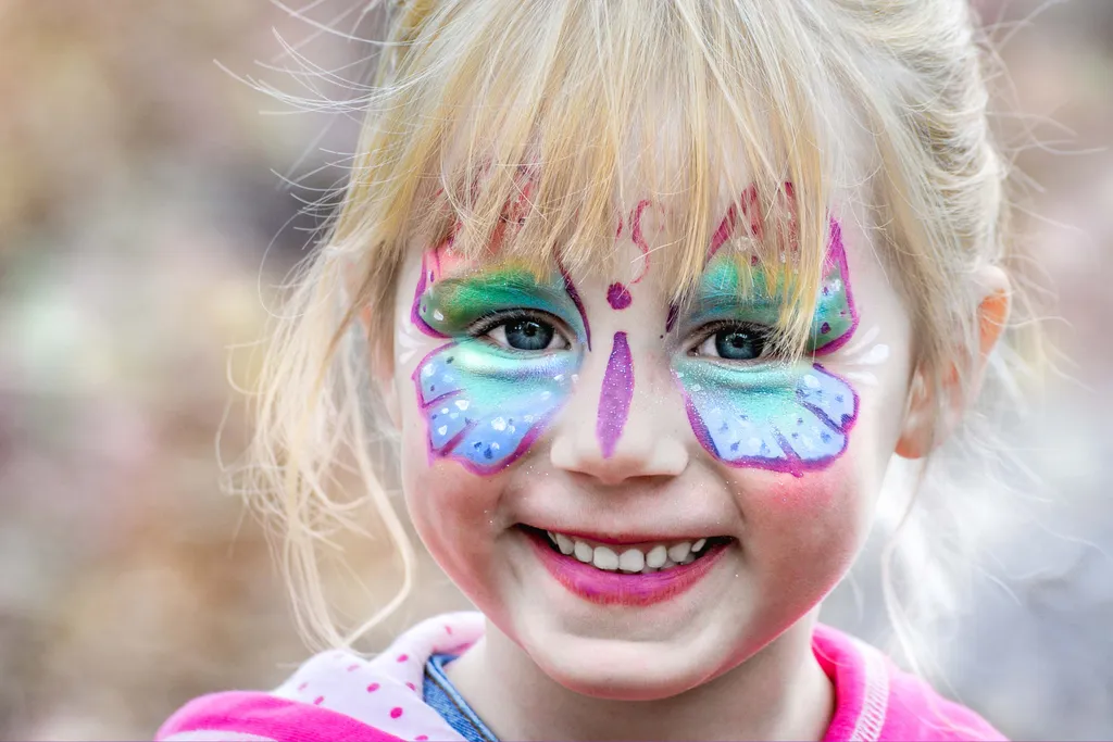 Smiling child with colorful butterfly face paint, featuring vibrant blues, greens, and pinks, and a bright pink shirt.
