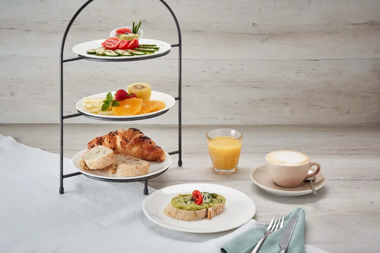 Ein dreistöckiger Ständer mit Obst, Gemüse, Brot und Croissants, dazu Orangensaft und ein Cappuccino auf einem Holztisch.