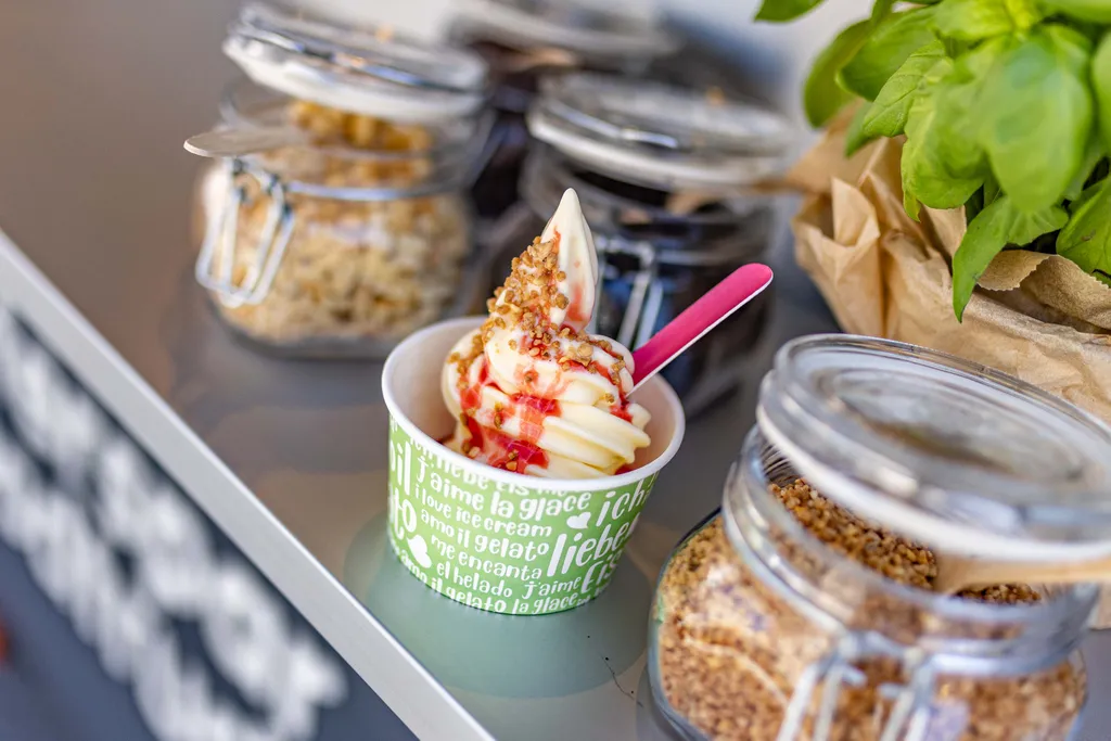 Softeis in einem grünen Becher mit rosa Löffel, garniert mit Nüssen und Erdbeersauce. Im Hintergrund stehen Gläser mit Toppings.