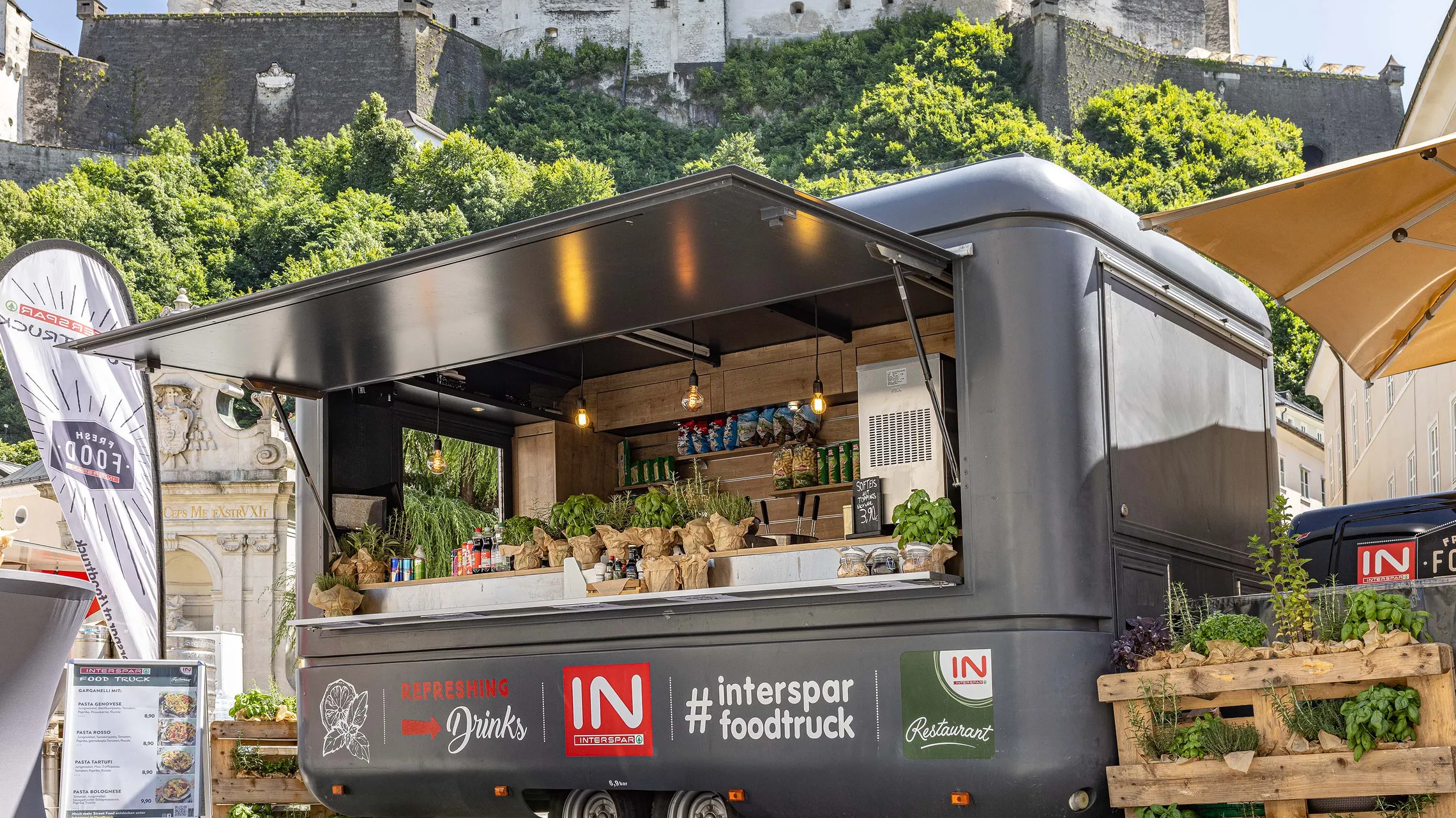 Ein Food Truck im Freien, an dem frische Produkte, Snacks und Getränke verkauft werden, vor dem Hintergrund der Festung Hohen Salzburg.