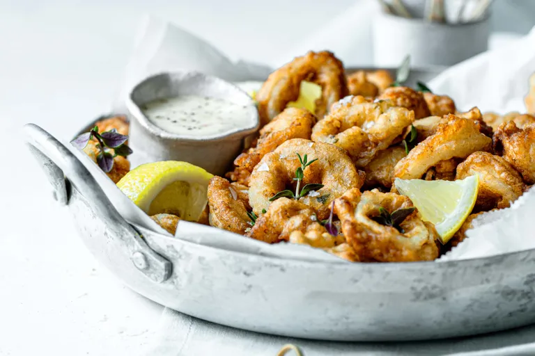 Ein Teller mit knusprigen Calamari Fritti, serviert mit Zitronenschnitzen und einer Dip-Sauce in einer rustikalen Metallschale, garniert mit Kräutern.