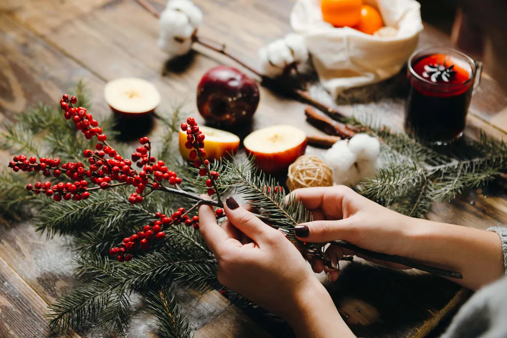 Hände, die rote Beeren und Fichtenzweige auf einem Tisch mit Apfelscheiben, Watte und einem Glas Glühwein arrangieren.