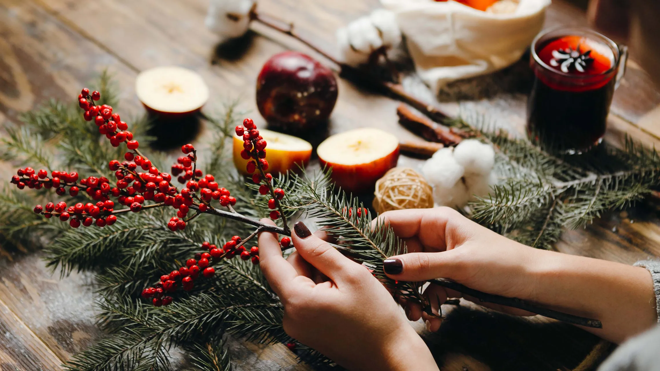 Hände, die rote Beeren und Fichtenzweige auf einem Tisch mit Apfelscheiben, Watte und einem Glas Glühwein arrangieren.