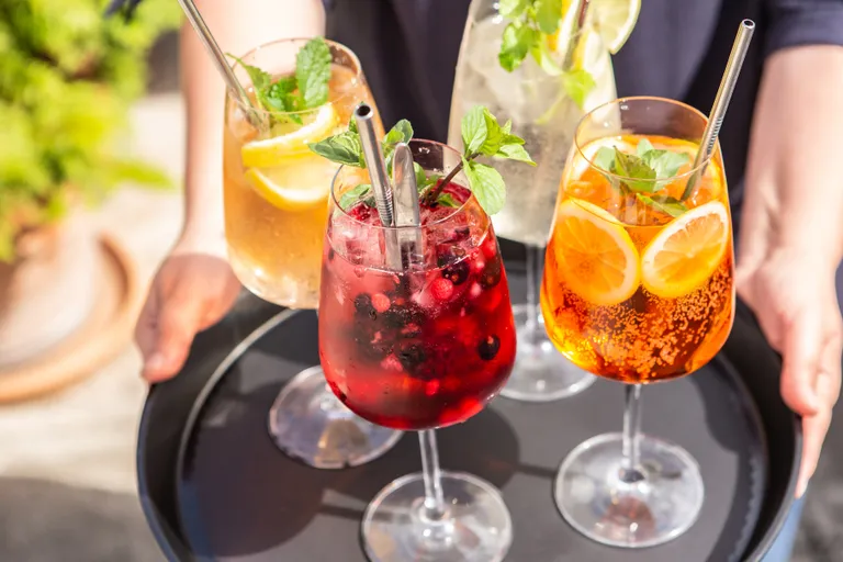 Ein Tablett mit vier bunten Cocktails, garniert mit Früchten und Minze, serviert an einem sonnigen Tag in klaren Gläsern mit Metallstrohhalmen.