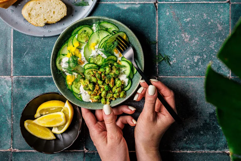 Hände, die eine Schüssel Gurkensalat mit Erbsen, Kräutern und Dressing halten. Daneben liegen Brotscheiben und Zitronenschnitze auf einer blaugrünen Fliesenoberfläche.