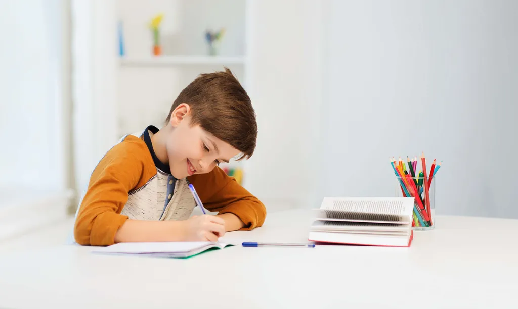 Junge schreibt konzentriert Hausaufgaben an einem Tisch mit Büchern und Buntstiften