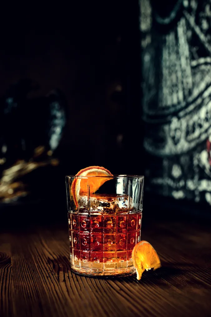 Ein Cocktailglas mit einem Eiswürfel und einer Orangenscheibe, das auf einem Holztisch steht, vor einem dunklen, unscharfen Hintergrund.