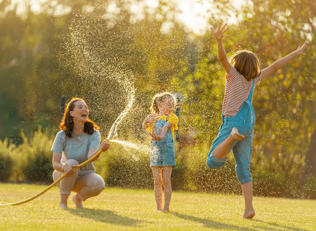 Eine Frau und zwei Kinder spielen fröhlich mit einem Gartenschlauch und spritzen Wasser in einem sonnendurchfluteten Garten.