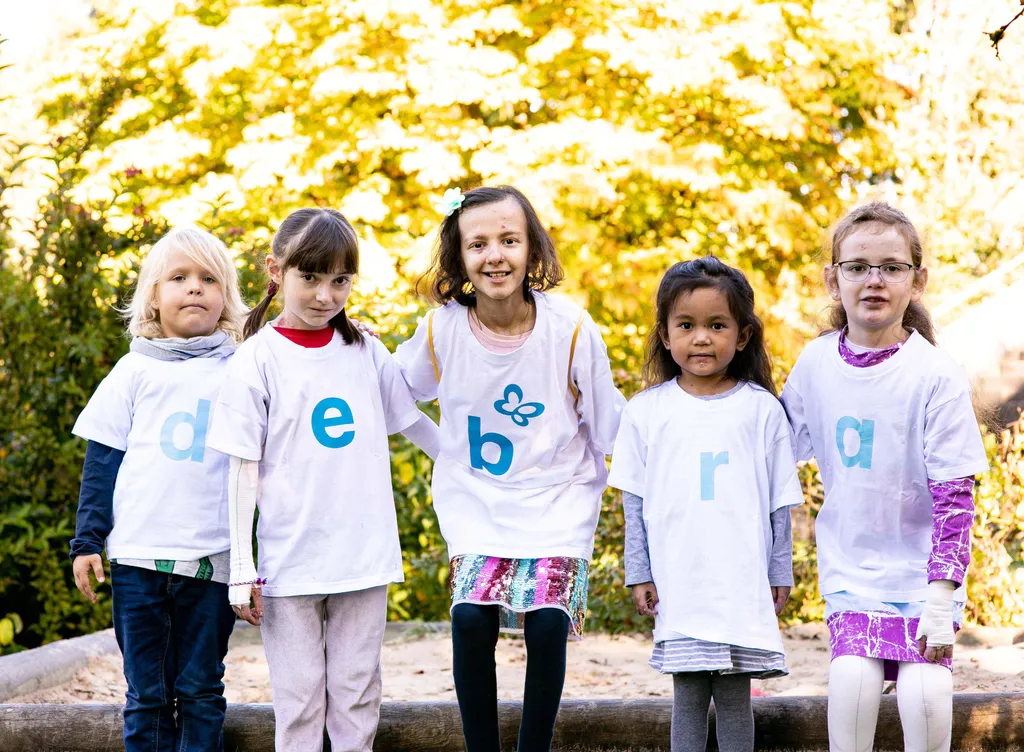 Mehrere Kinder mit weißen T-Shirts auf denen sich jeweils ein Buchstabe befindet, die zusammen das Wort "debra" ergeben