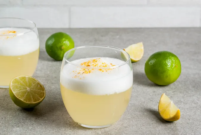 Ein schaumiger Cocktail in einem Glas, garniert mit Orangenschale, umgeben von ganzen und in Scheiben geschnittenen Limetten auf einer grauen Unterlage.