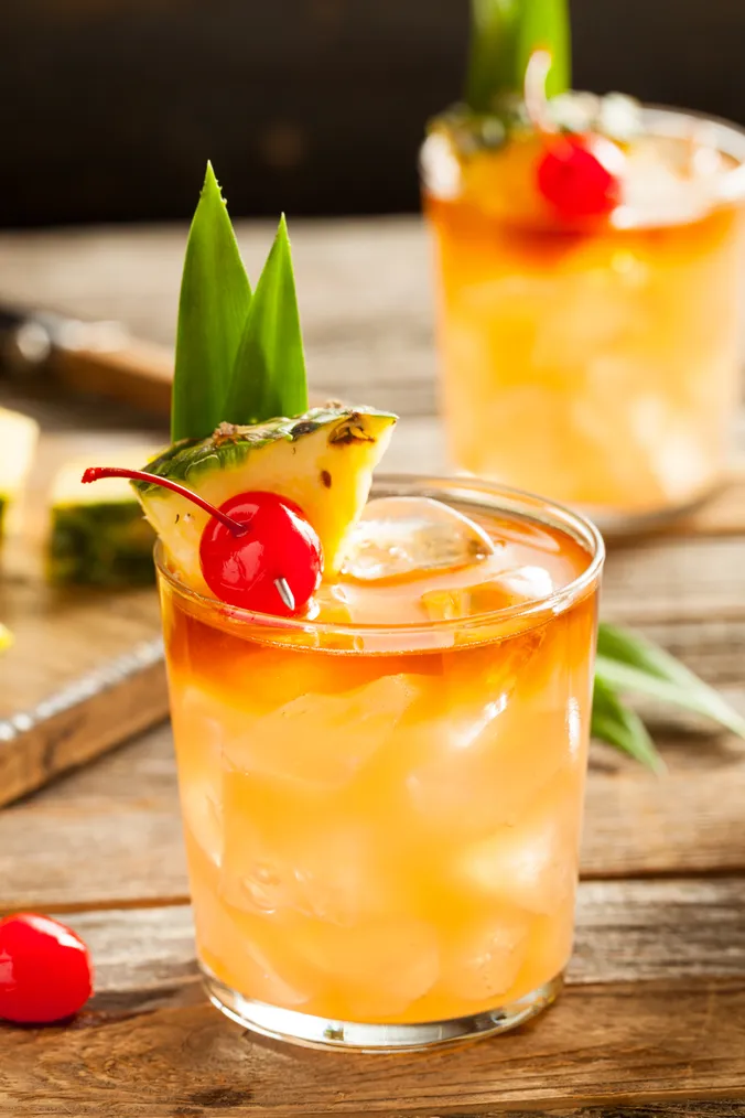 Ein erfrischender tropischer Cocktail mit Ananas, Kirsche und Blattgarnitur in einem mit Eis gefüllten Glas, das auf einem Holztisch steht.