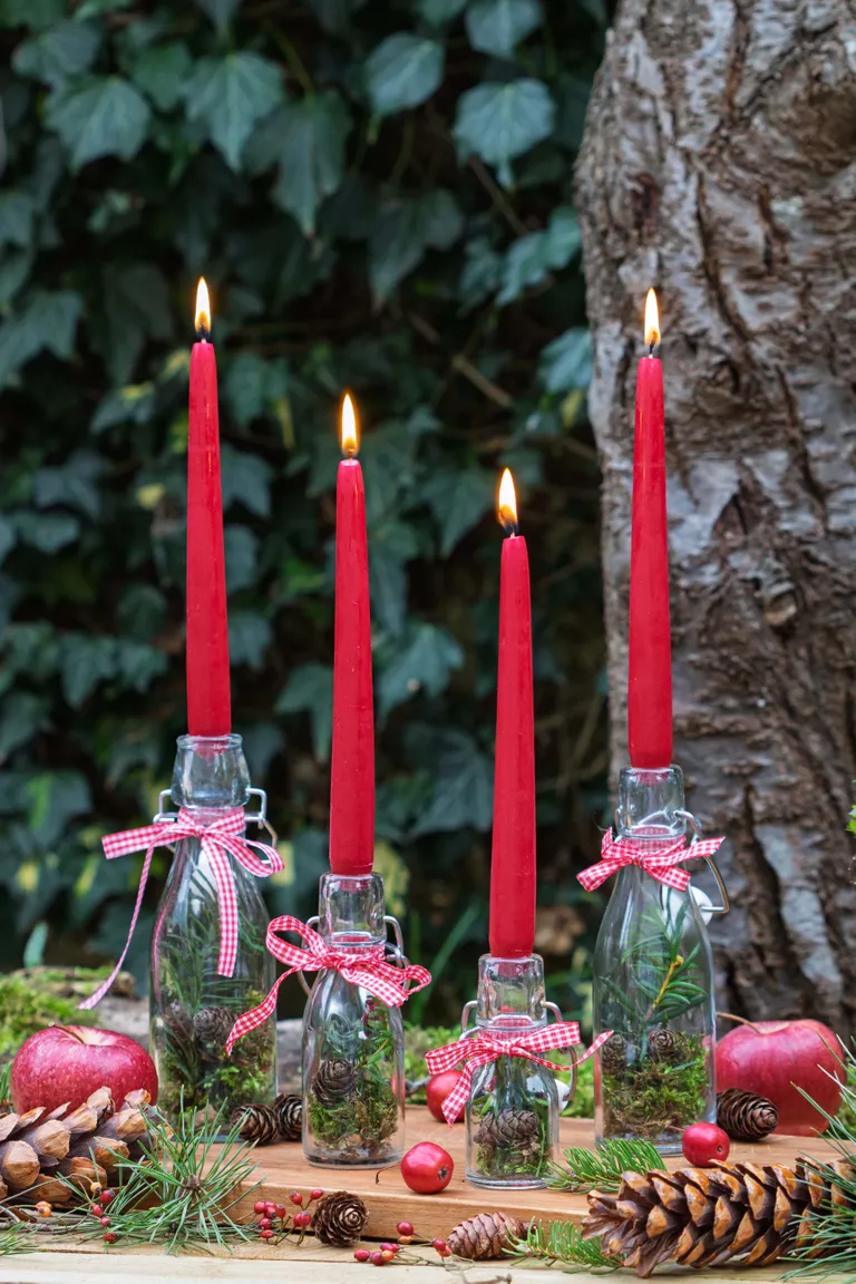 Vier rote Kerzen in Glasflaschen mit roten Bändern, umgeben von Tannenzapfen, Äpfeln und Beeren, vor einem Hintergrund aus Blättern.