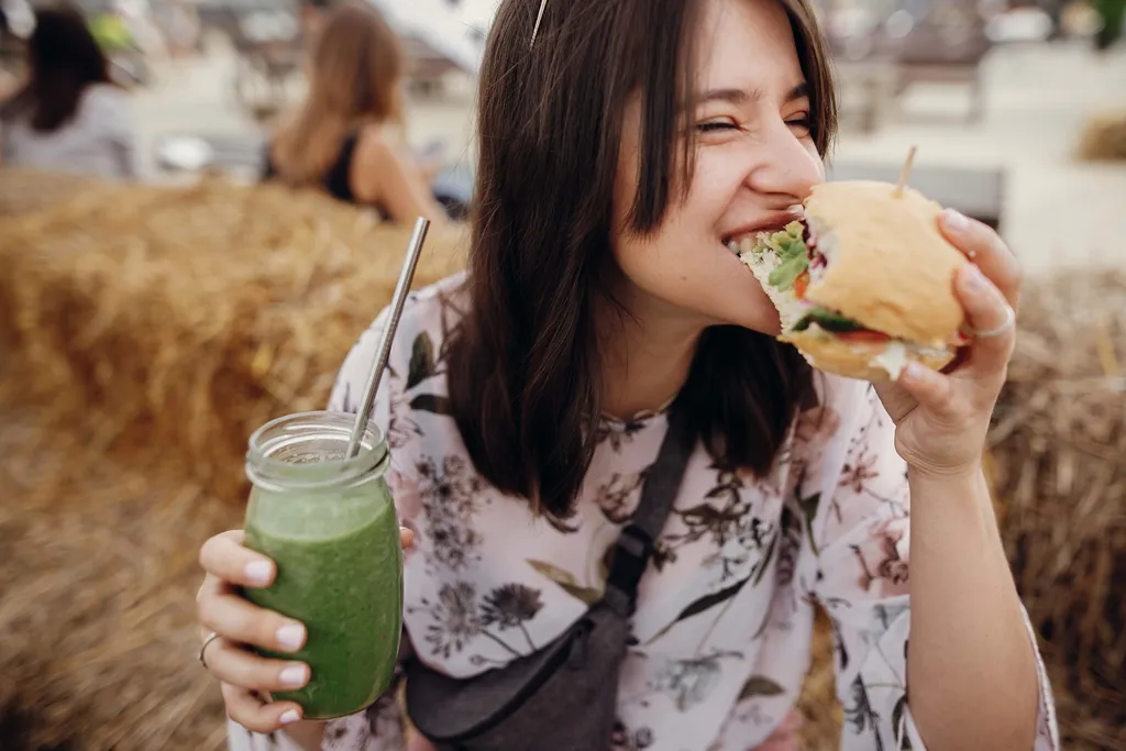 Eine Frau in einer geblümten Bluse genießt einen Burger und einen grünen Smoothie, während sie auf einem Heuballen sitzt.
