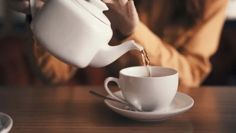 Eine Person beim Einschenken von frischem Tee aus einer weißen Teekanne in eine weiße Teetasse