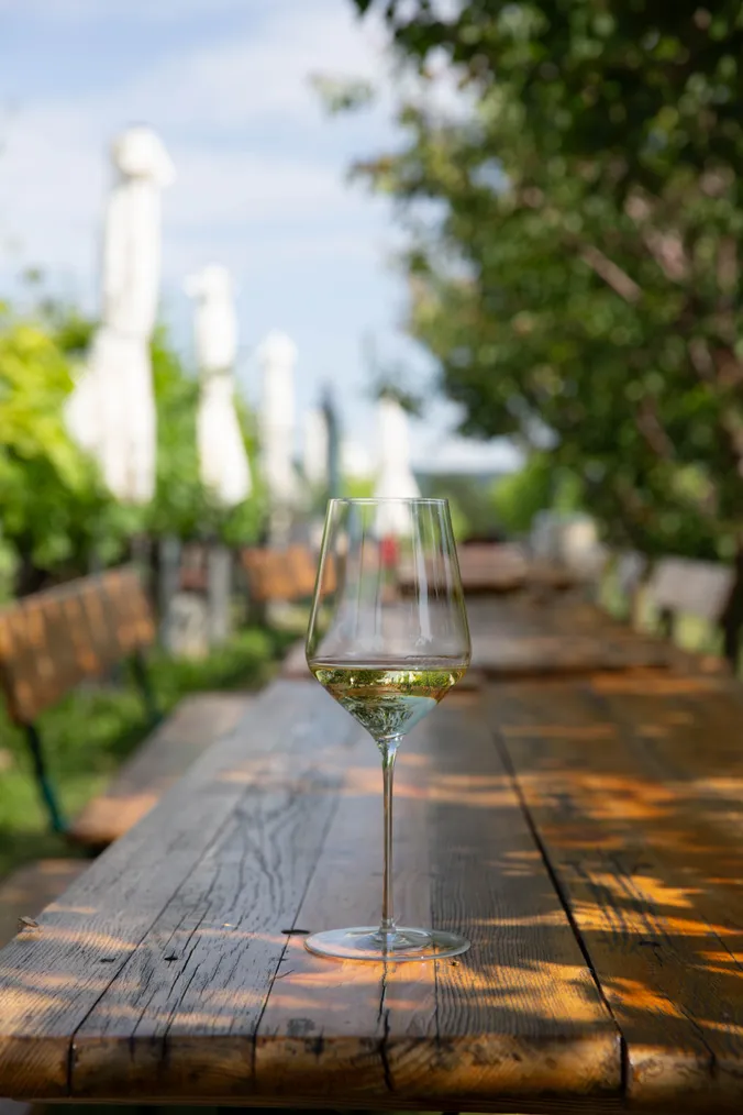 Ein Glas Weißwein auf einem rustikalen Holztisch in einer sonnigen Außenkulisse mit viel Grün und unscharfen Sonnenschirmen im Hintergrund.