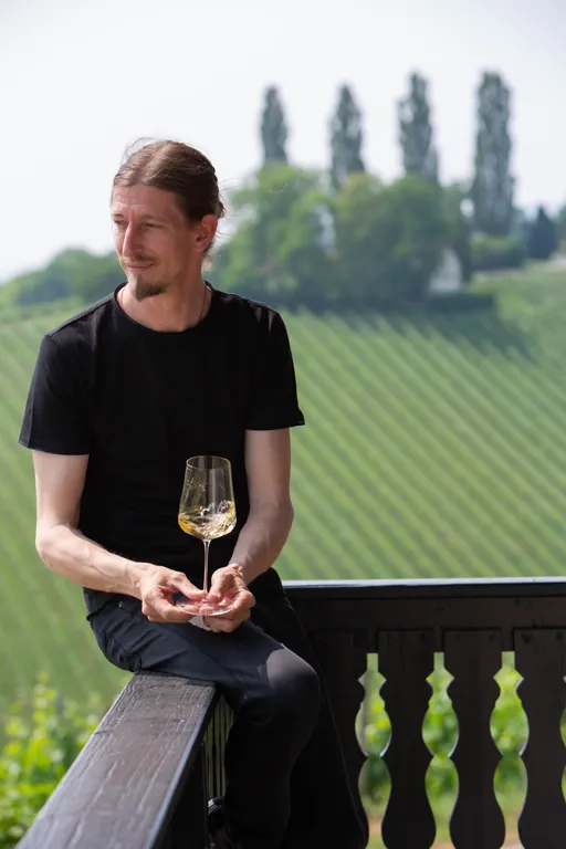 A man in a black shirt sits on a railing, holding a wine glass, with a scenic vineyard and trees in the background.