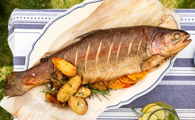 Ein ganzer gegrillter Fisch auf einer Platte mit Bratkartoffeln, Orangenscheiben und Rosmarin, serviert auf einer gestreiften Tischdecke.