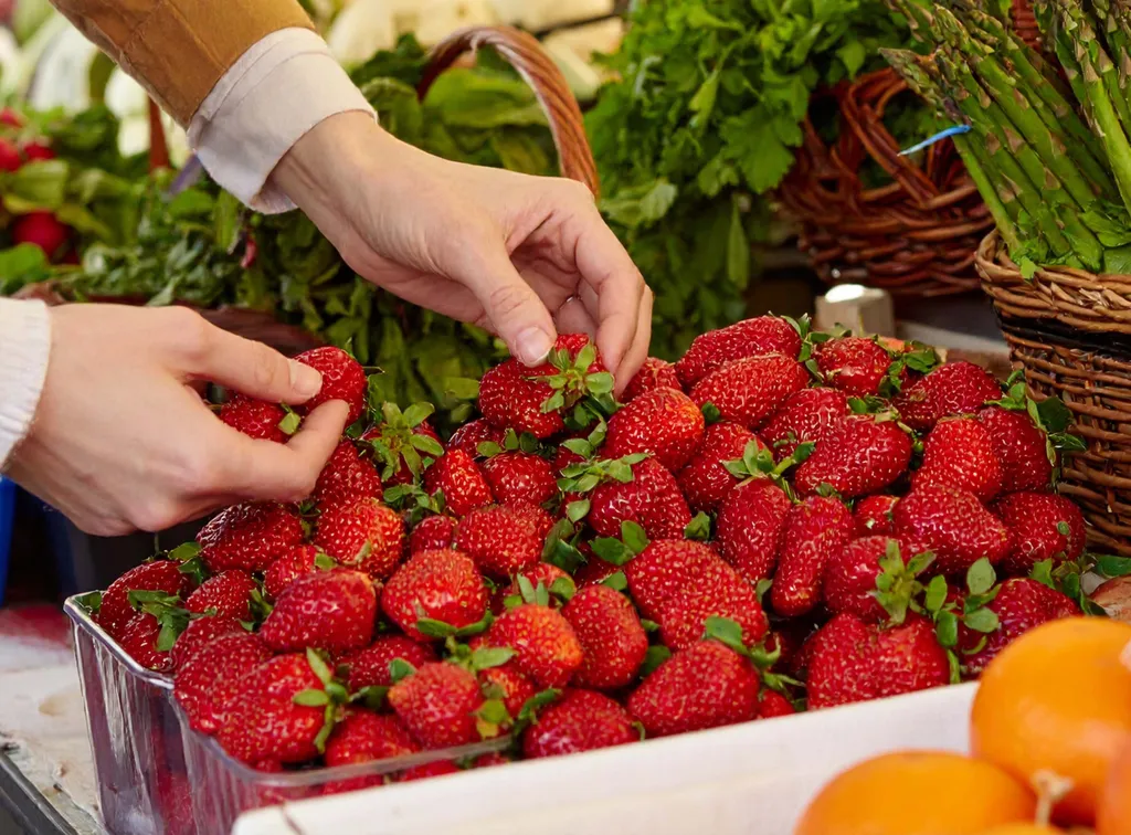Hände, die auf einem Markt frische Erdbeeren aus einem Haufen aussuchen, umgeben von grünem Gemüse und Orangen.