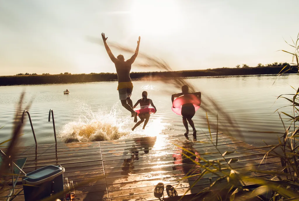 Drei Menschen springen bei Sonnenuntergang fröhlich von einem Steg in einen See, wobei einer eine rosa Schwimmhilfe benutzt und dabei Spritzer im Wasser verursacht.