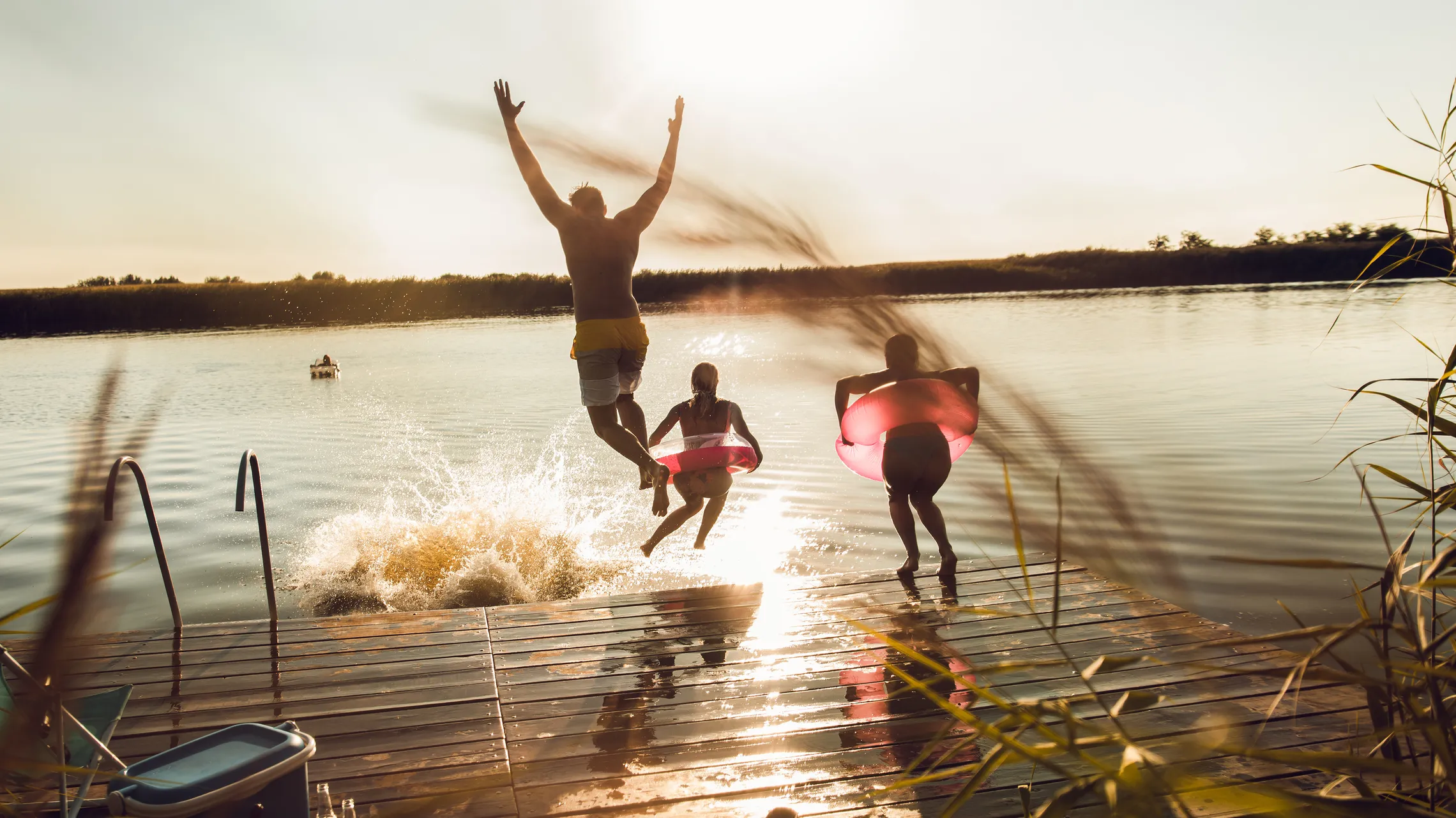 Drei Menschen springen bei Sonnenuntergang fröhlich von einem Steg in einen See, wobei einer eine rosa Schwimmhilfe benutzt und dabei Spritzer im Wasser verursacht.