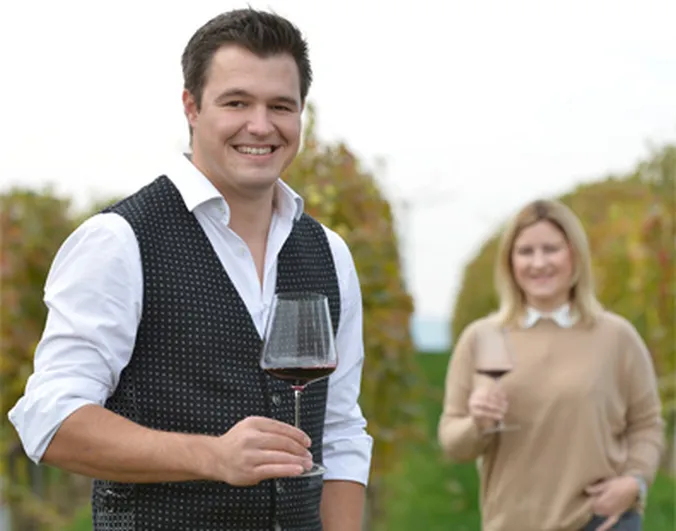 Ein Mann und eine Frau stehen in einem Weinberg, beide halten ein Glas Rotwein in der Hand und lächeln.