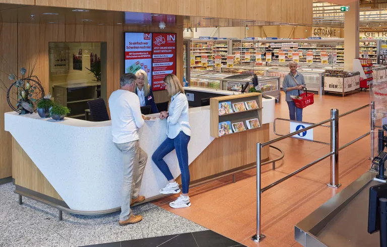 Empfangsbereich in einem Geschäft, in dem zwei Kunden mit der Empfangsmitarbeiterin sprechen. Im Hintergrund sind Regale mit Produkten zu sehen.