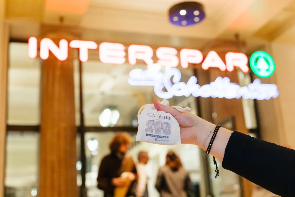Eine Hand hält ein Säckchen Genussgulden vor das Eingangsschild INTERSPAR am Schottentor