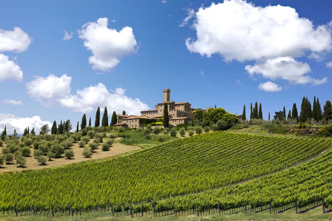 Eine toskanische Villa auf einem Hügel mit Zypressen und Weinbergen unter blauem Himmel mit flauschigen Wolken.