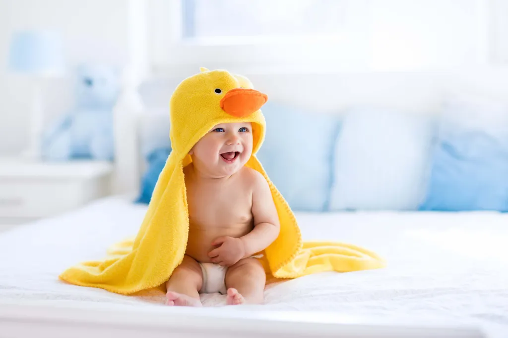 Lachendes Baby sitzt mit gelbem Kapuzenhandtuch auf dem Bett