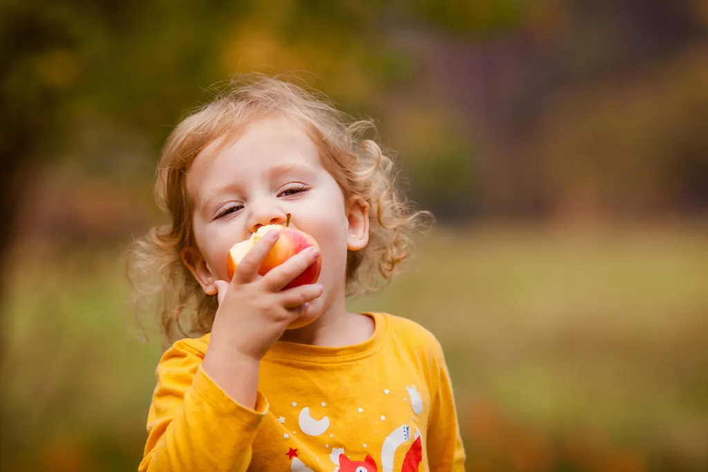 Ein Kind mit lockigem Haar genießt einen Bissen von einem roten Apfel; es trägt ein gelbes Hemd und steht im Freien vor einem verschwommenen grünen Hintergrund.