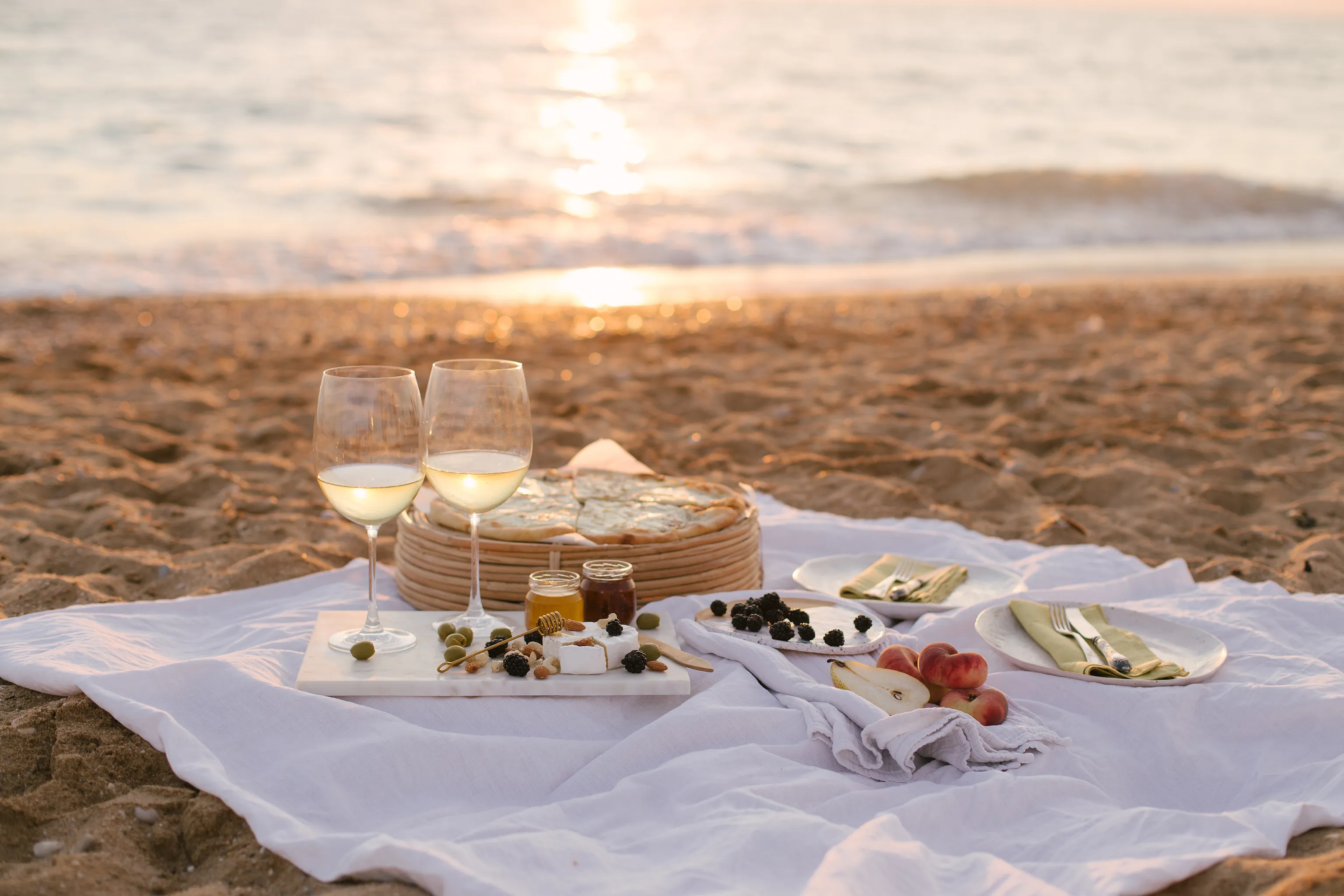 Ein Picknick am Strand bei Sonnenuntergang mit zwei Gläsern Weißwein, Obst, Käse, Oliven und Brot auf einer weißen Tischdecke, mit dem Meer im Hintergrund.
