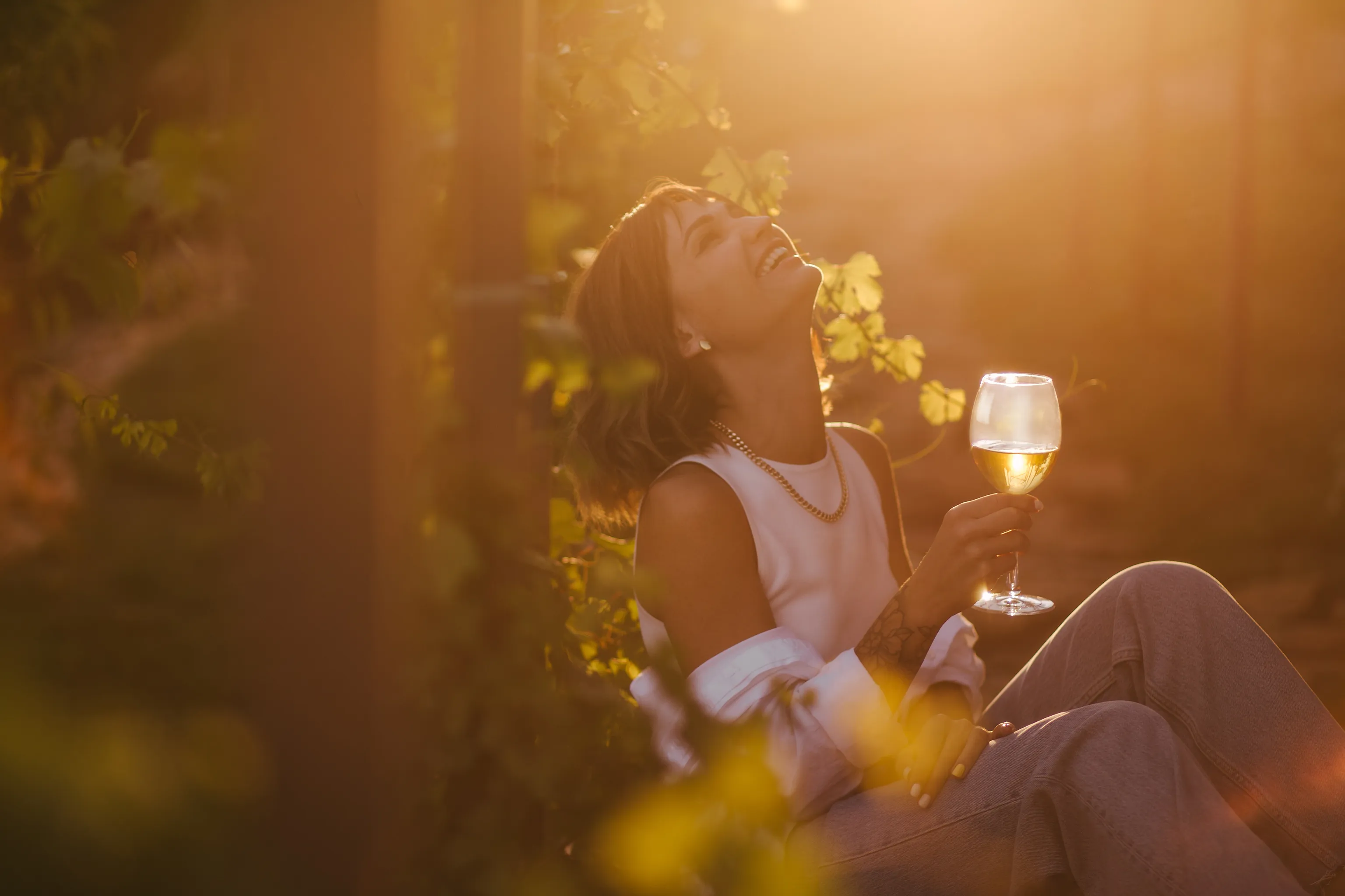 Eine Frau sitzt in einem Weinberg, lacht und hält ein Glas Weißwein in der Hand, beleuchtet von warmem, goldenem Sonnenlicht.