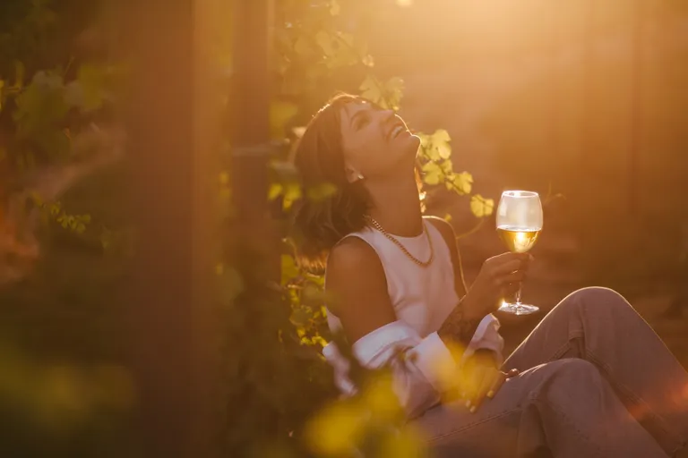 Eine Frau sitzt in einem Weinberg, lacht und hält ein Glas Weißwein in der Hand, beleuchtet von warmem, goldenem Sonnenlicht.