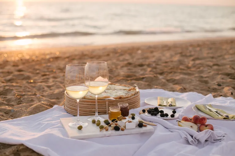 Ein Picknick am Strand mit zwei Gläsern Weißwein, einer Käseplatte, Oliven, Obst und Brot auf einer weißen Decke bei Sonnenuntergang.