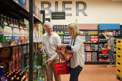 Zwei Menschen, die lächelnd in einem Supermarkt Bier aus den Regalen aussuchen. Im Hintergrund ist eine weitere Person beim Einkaufen zu sehen.