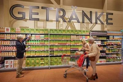 Drei Personen, die in einem Getränkegang voller bunter Getränkverpackungen unter einem großen „GETRÄNKE“-Schild einkaufen.