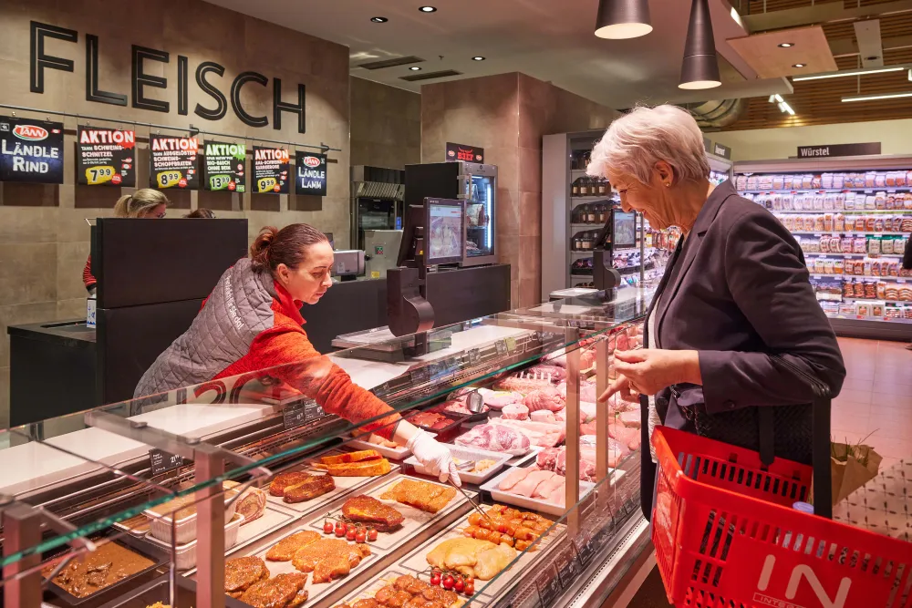 Eine Frau wählt an einer Fleischtheke unter Anleitung eines Metzgers Produkte aus, während unter einem „Fleisch“-Schild in einem Supermarkt verschiedene Fleischsorten ausgestellt sind.