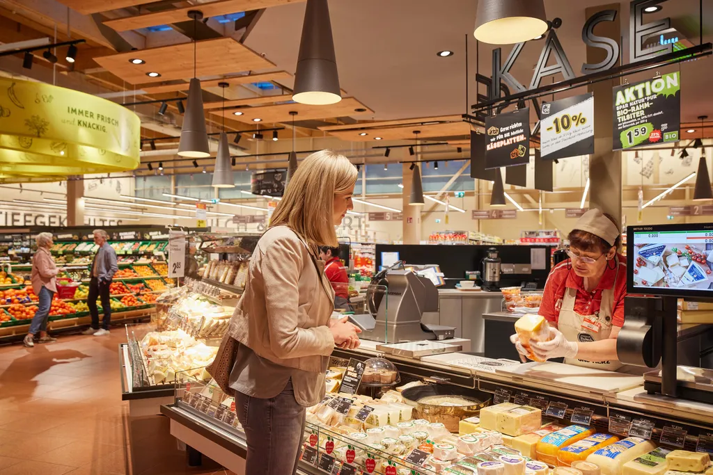 Eine Frau an der Käsetheke eines Supermarkts, die von einem Mitarbeiter bedient wird, im Hintergrund Regale mit Lebensmitteln und andere Kunden.