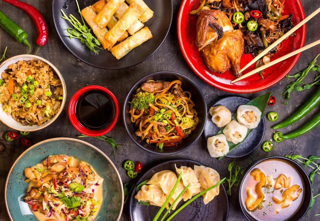 Eine Auswahl an asiatischen Gerichten, darunter Nudeln, Teigtaschen, Frühlingsrollen, gebratener Reis, Brathähnchen und Suppe, garniert mit Chilischoten und Kräutern.