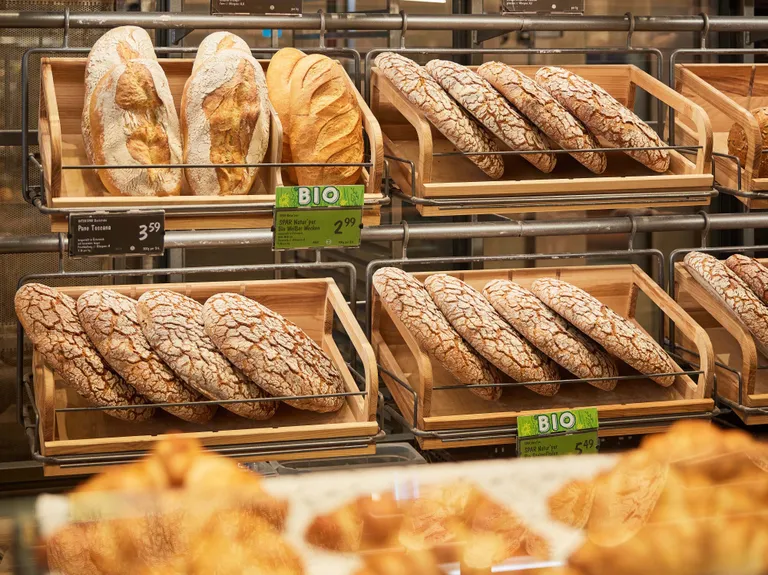 Auf Holztabletts liegen in einer Bäckerei verschiedene handwerklich hergestellte Brotsorten, darunter Laibe und Rundbrote, mit gut sichtbaren Preisschildern.