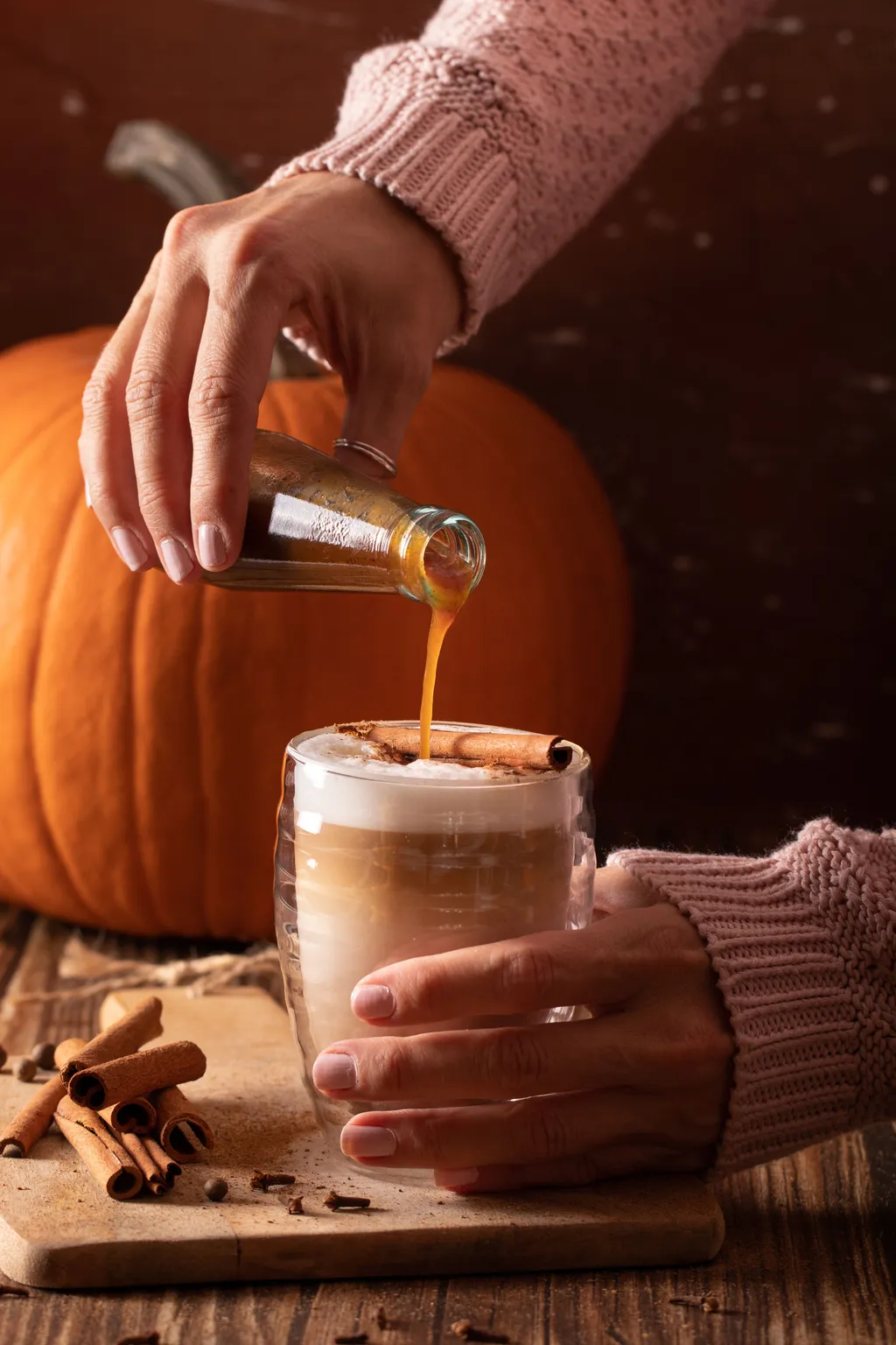 Die Händer einer Frau in einem altrosa Strickpullover gießen Pumpkin Spice Sirup in ein Glas mit Milchkaffee, brauntoniger Hintergrund mit Kürbis