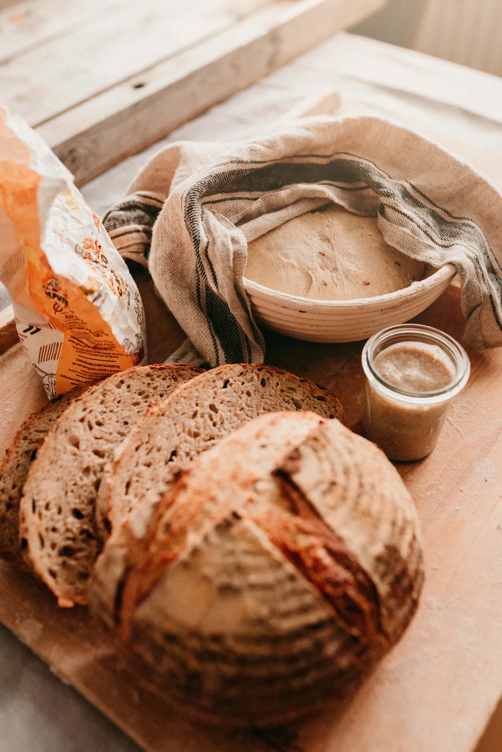 Frisch gebackenes Sauerteigbrot mit Brotscheiben, eine mit einem Tuch bedeckte Schüssel mit Teig, ein Mehlsack und ein Glas mit Sauerteigstarter auf einem Holzbrett.